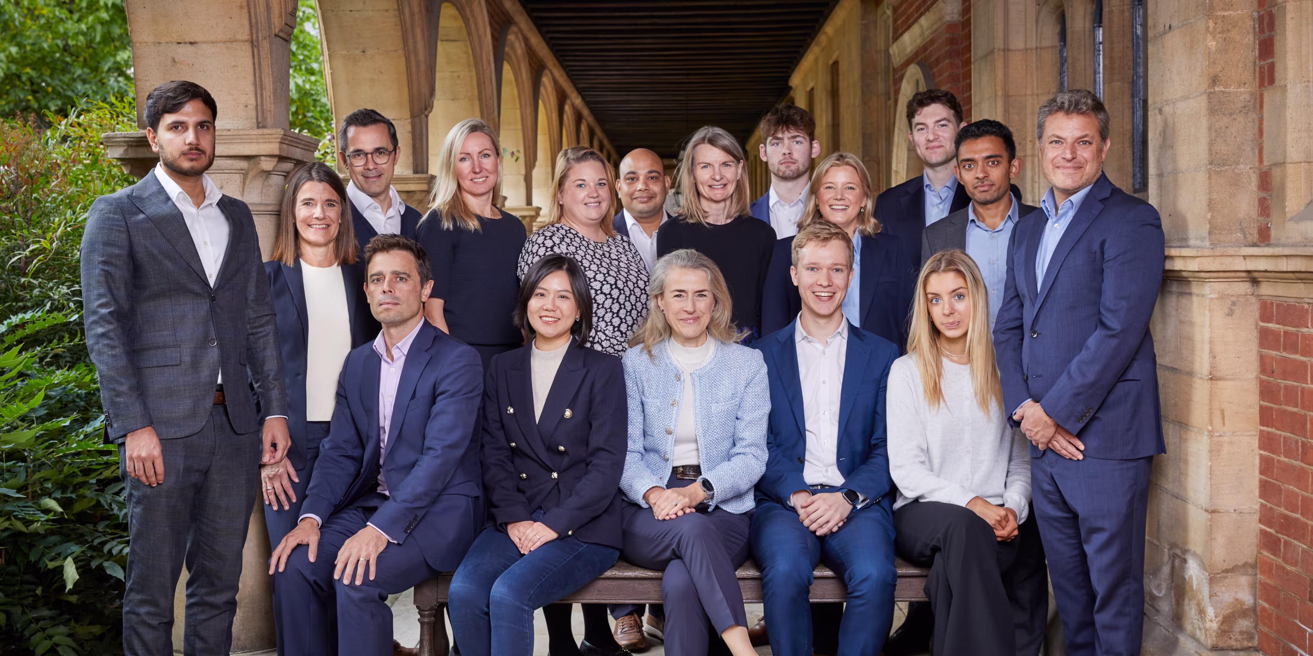 Group portrait of 16 professionally dressed diverse men and women posing in an outdoor arched walkway.