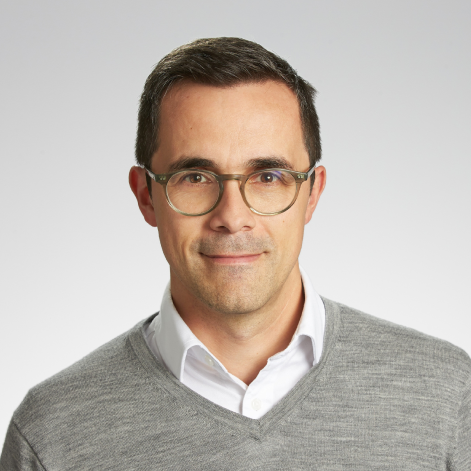 Man with short dark hair and glasses wearing a gray sweater over a white collared shirt against a light gray background.