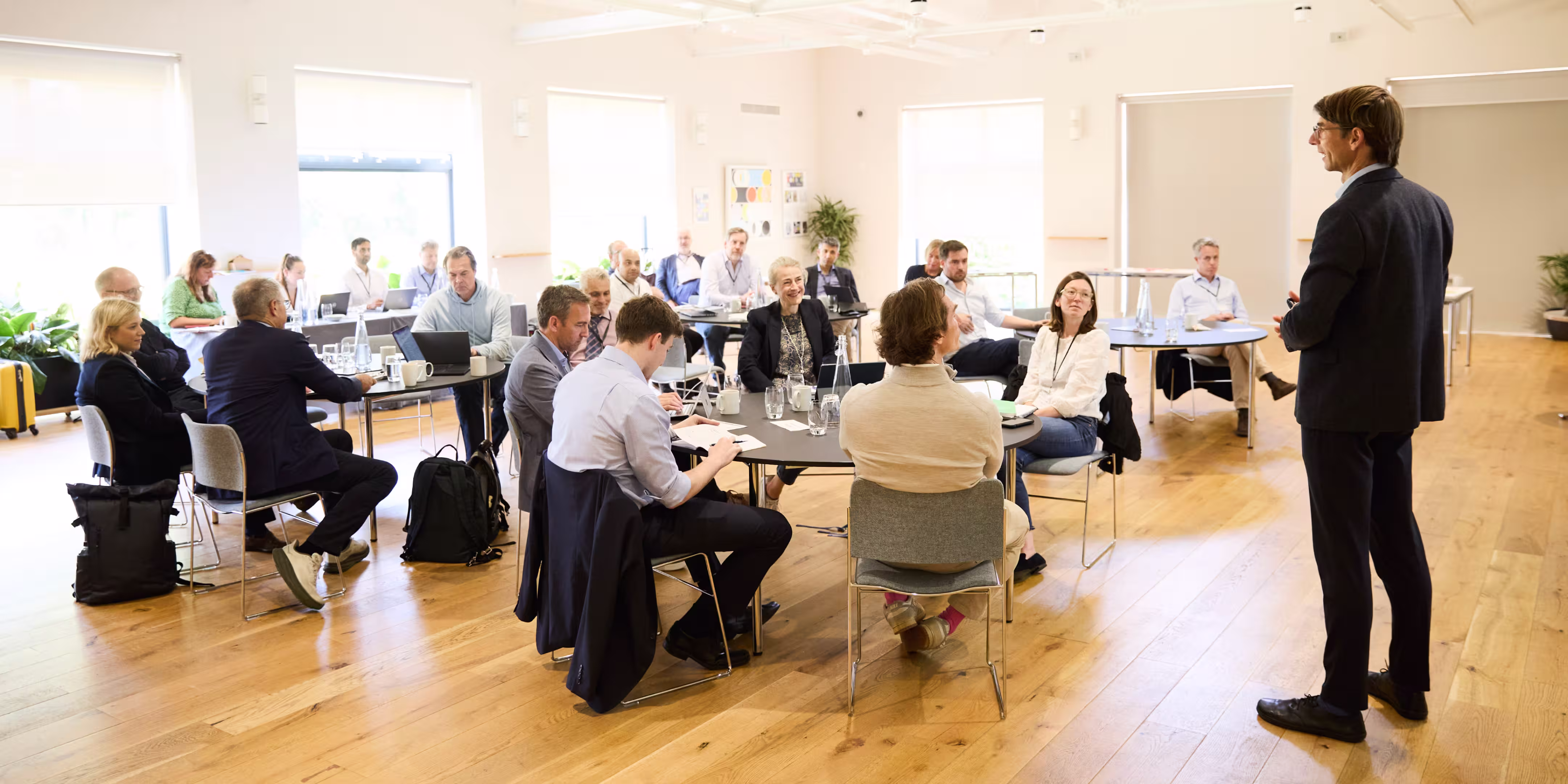 A professional speaker addresses a group of adults seated at round and rectangular tables in a bright, modern room during an executive education program.
