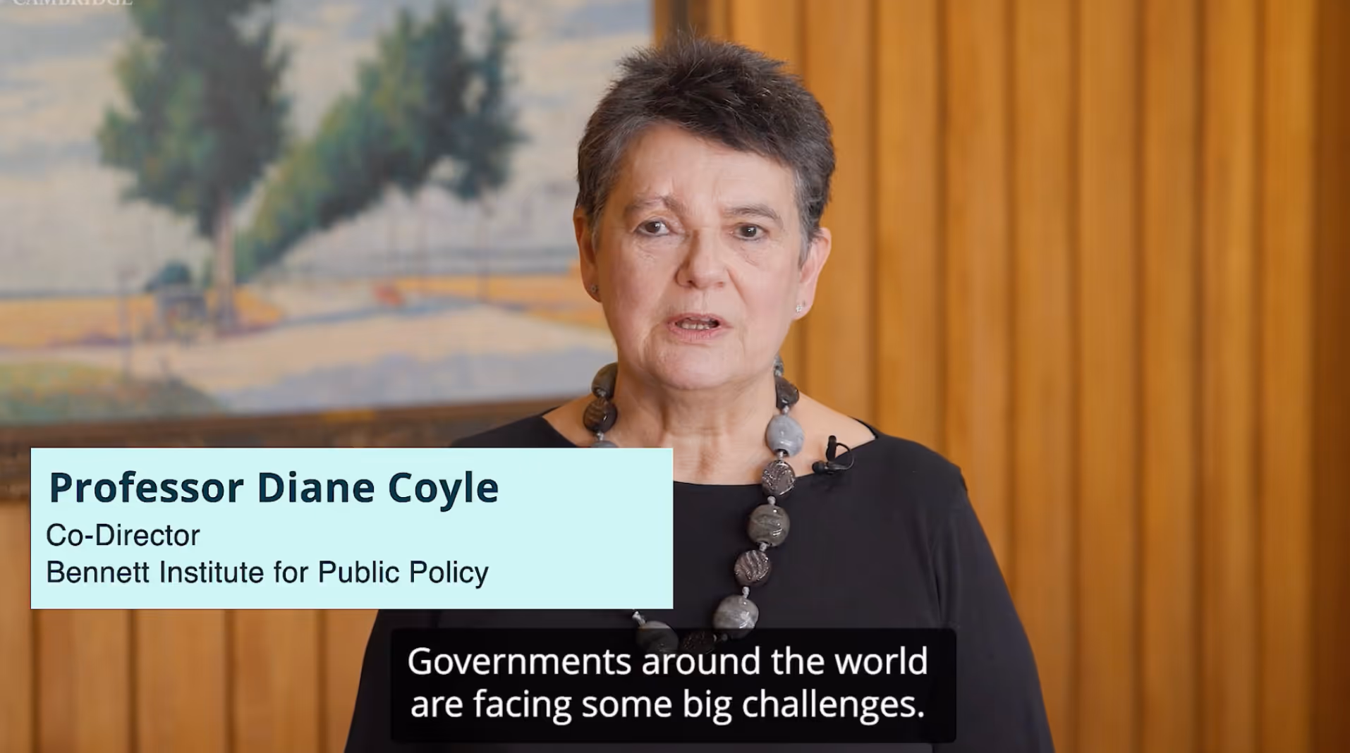 Professor Diane Coyle speaking in an indoor setting with a wooden panel background and text identifying her as Co-Director of Bennett Institute for Public Policy.