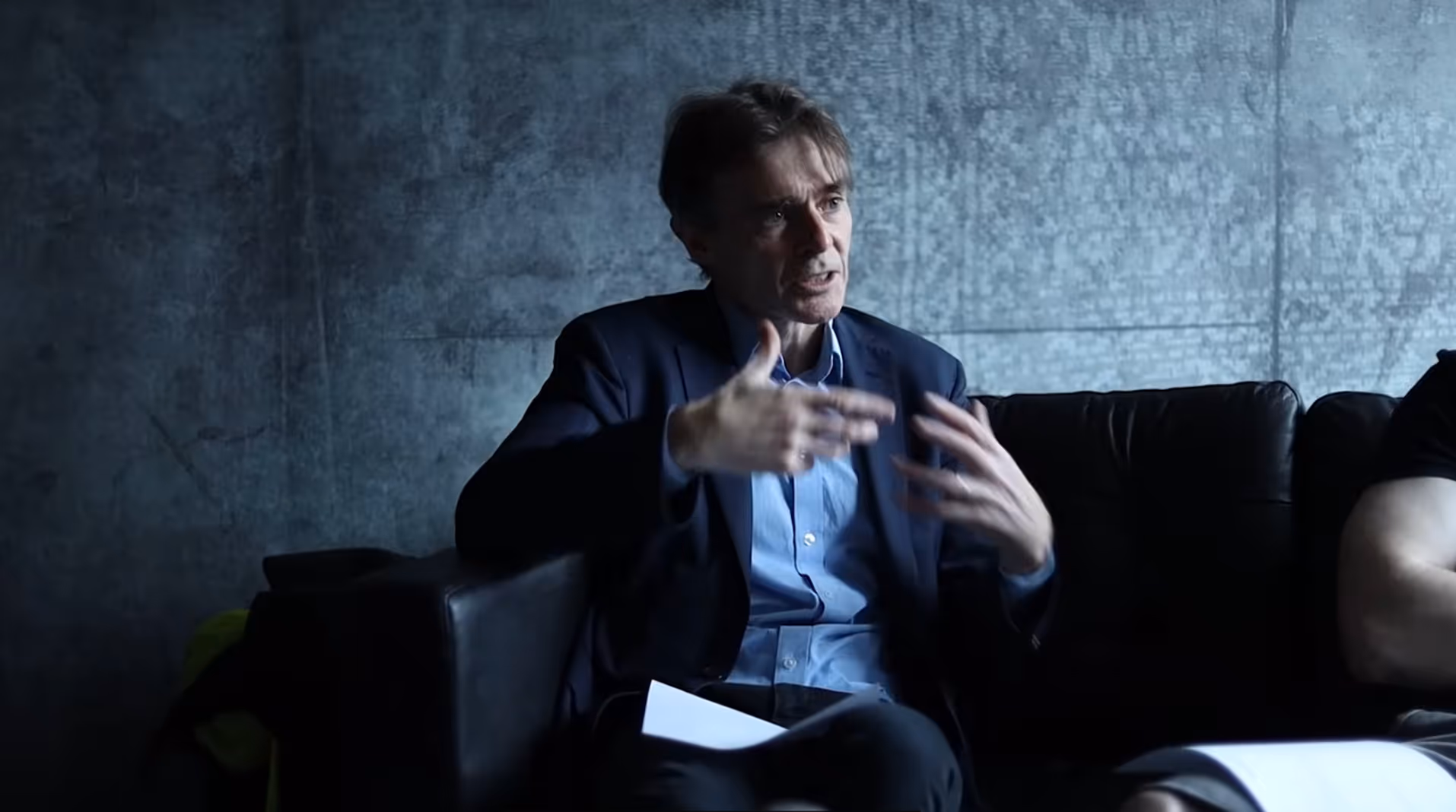 Man in a blazer and blue shirt sitting on a dark couch gesturing with his hands while holding papers.