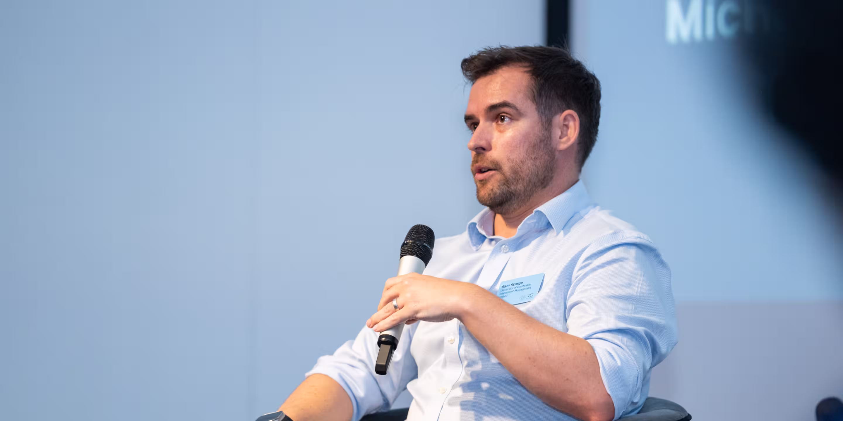 Man in light blue shirt speaking into a handheld microphone while seated.