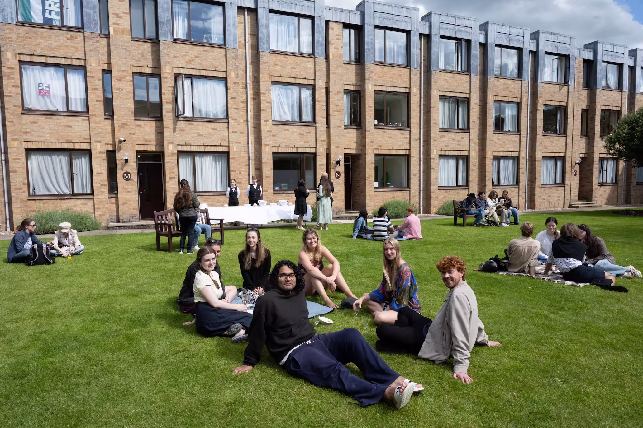 Groups of young adults sitting and socializing on a lawn in front of a brick building with several windows.