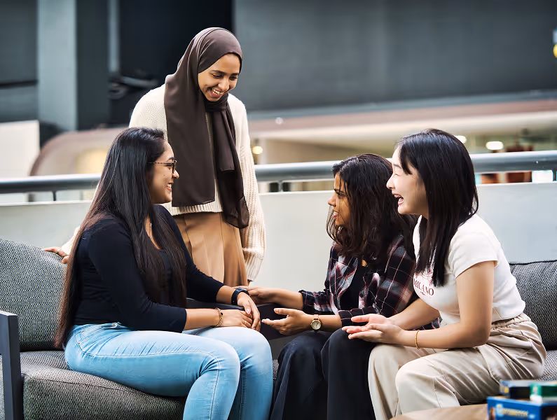 Four diverse women sitting and standing around a couch, engaging in a friendly conversation indoors.