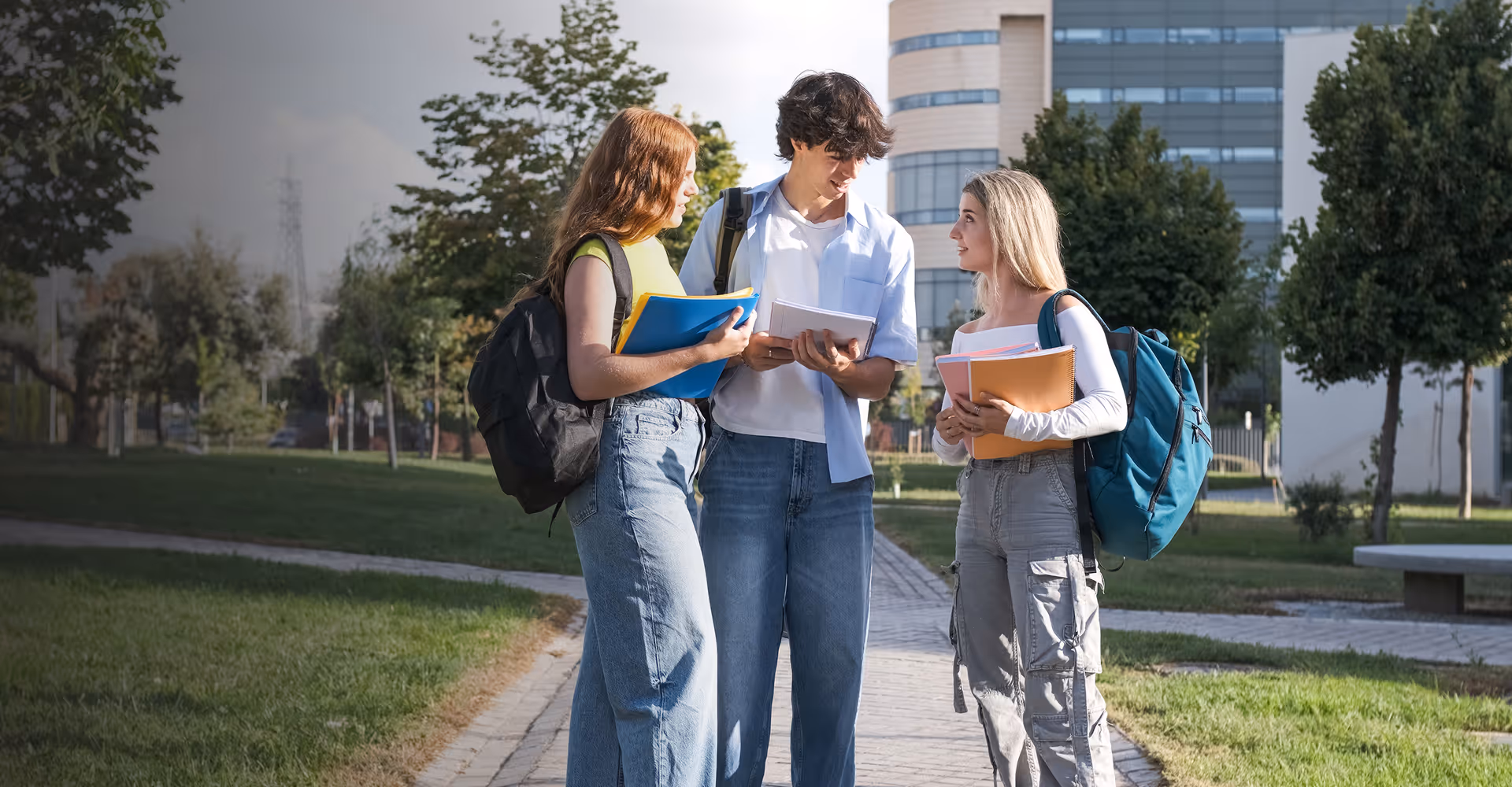 Three students standing outdoors on a campus pathway, holding notebooks and backpacks, engaged in conversation.