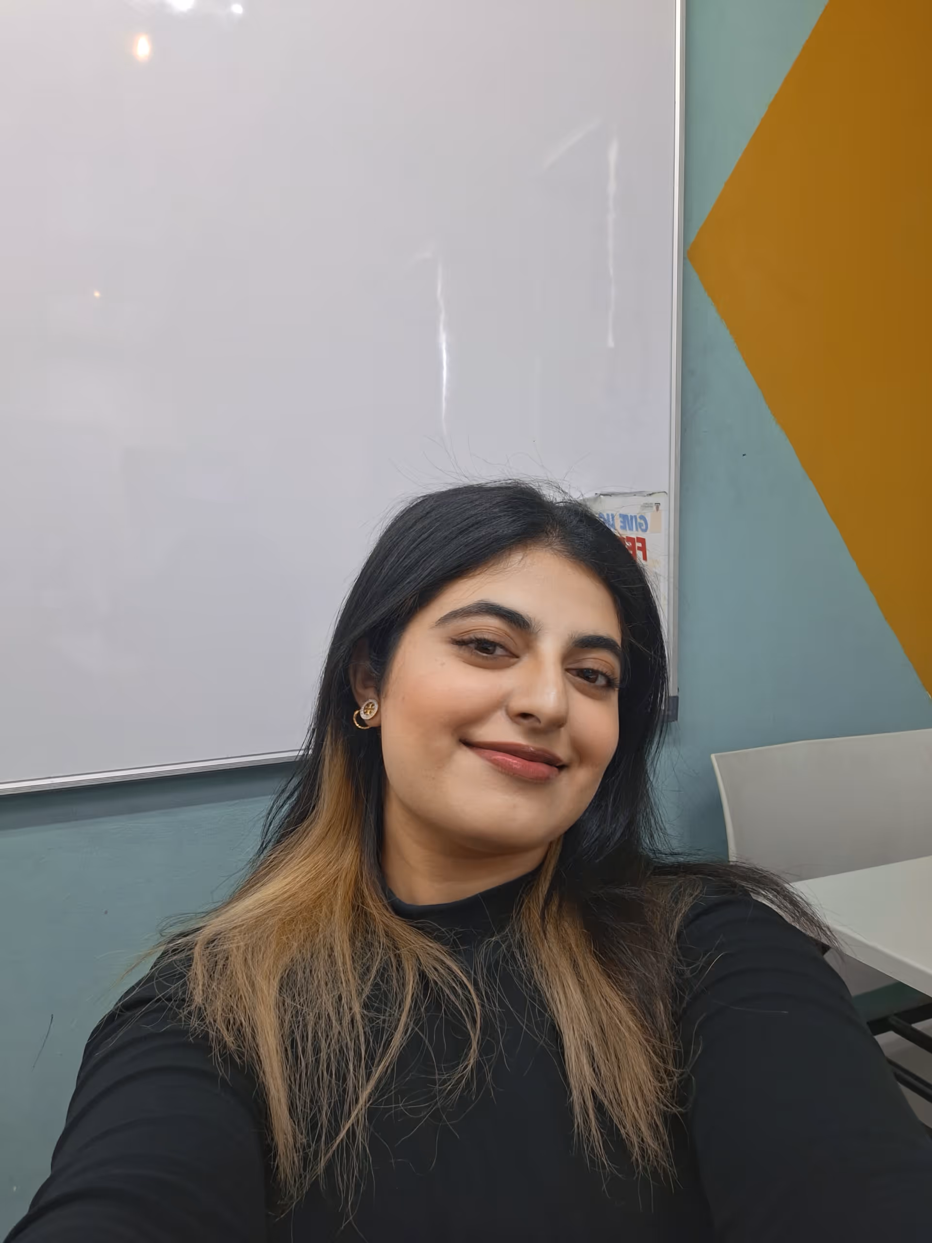 Smiling woman with long black and blonde hair wearing a black top, taking a selfie in a room with a whiteboard and geometric wall art.