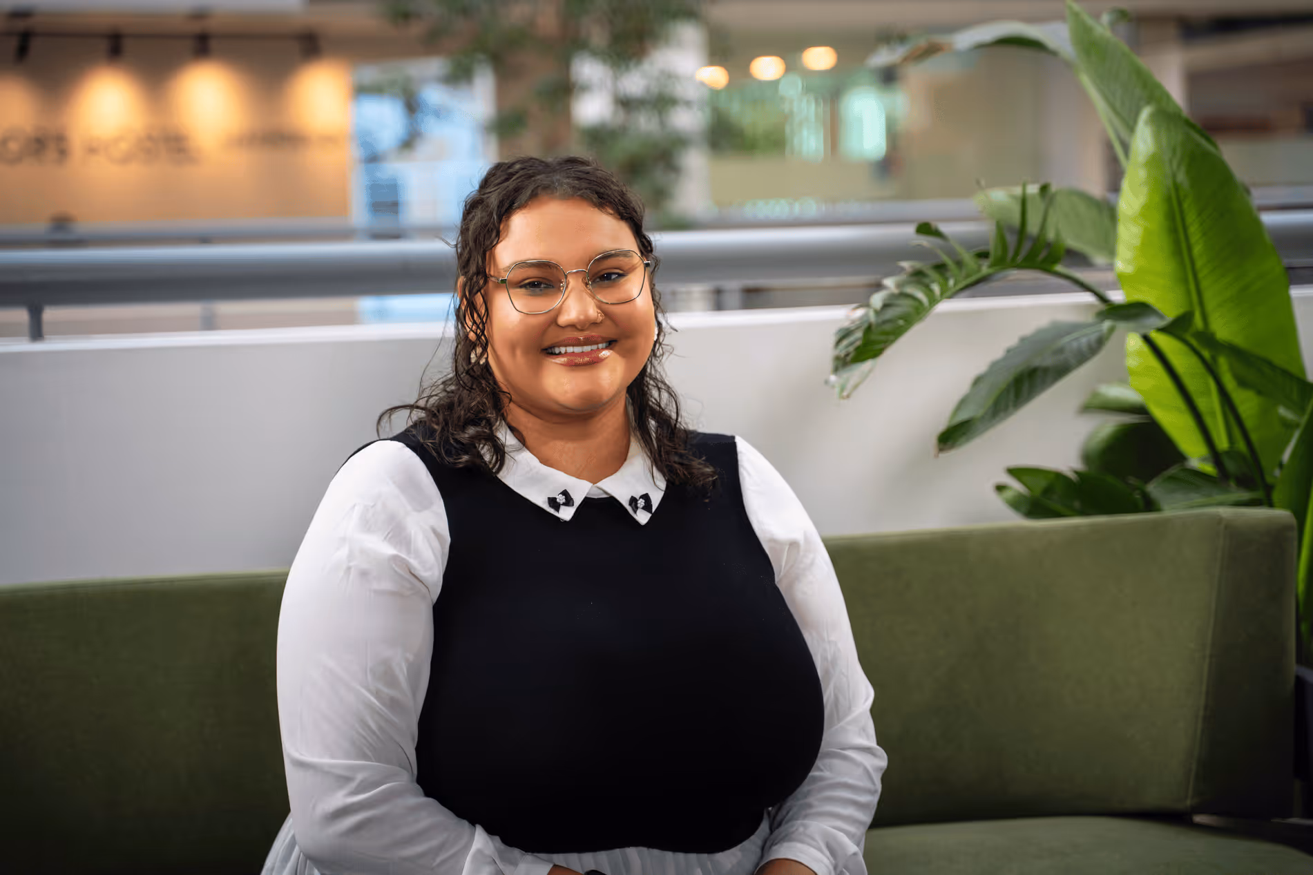 Smiling woman with glasses wearing a white collared shirt and black sleeveless sweater sitting on a green couch with plants in the background.