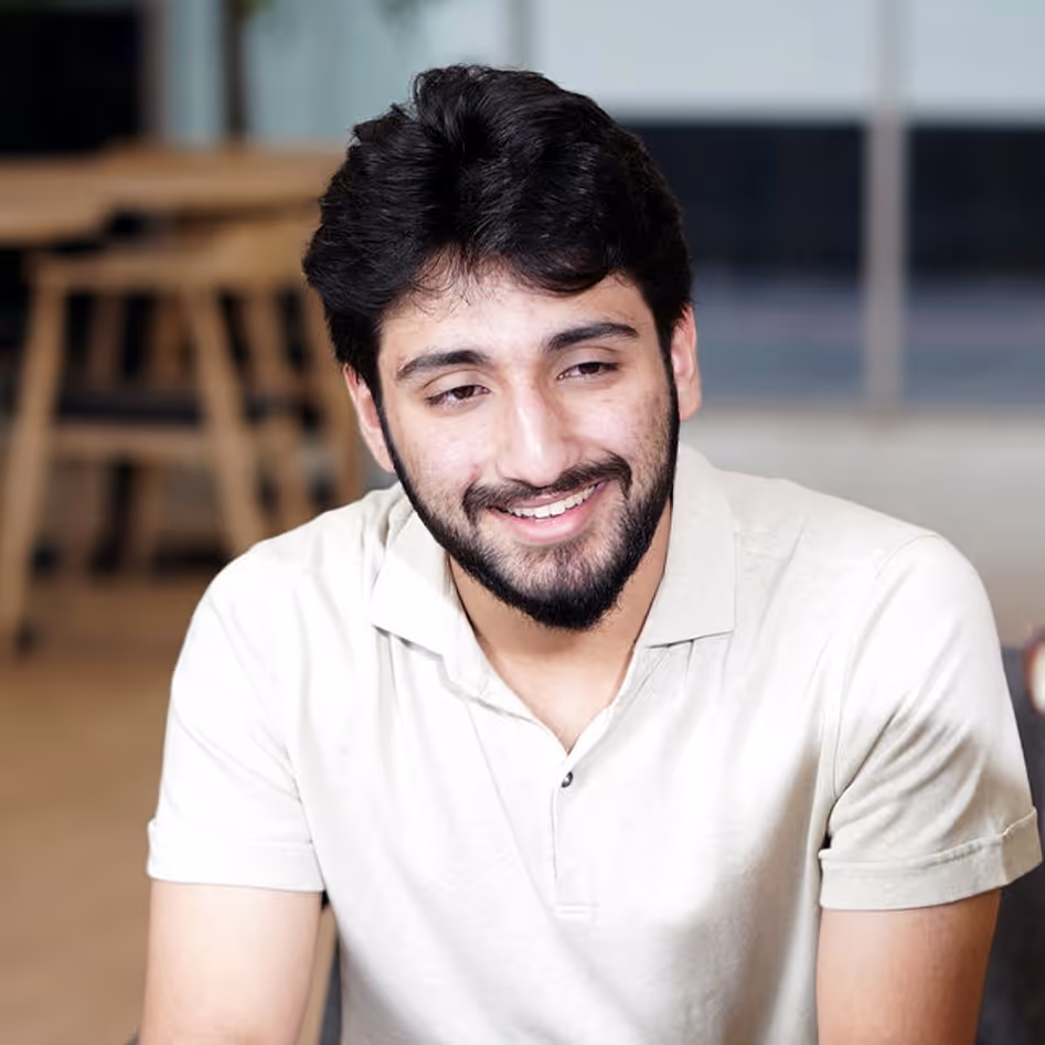 Smiling young man with short dark hair and beard wearing a light beige polo shirt in an indoor setting.