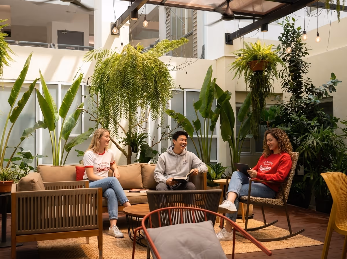 Three young adults sitting and chatting in a bright indoor patio surrounded by lush green plants and modern furniture.
