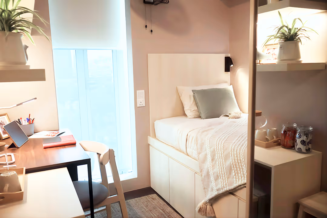 Cozy small bedroom with a single bed, light-colored bedding, a bedside shelf with plants and jars, and a wooden desk with a chair near a window.