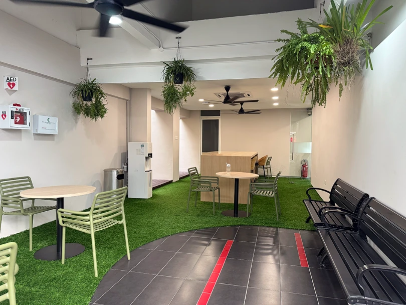 Indoor study area with artificial grass flooring, round tables with green chairs, black benches, hanging plants, and ceiling fans.