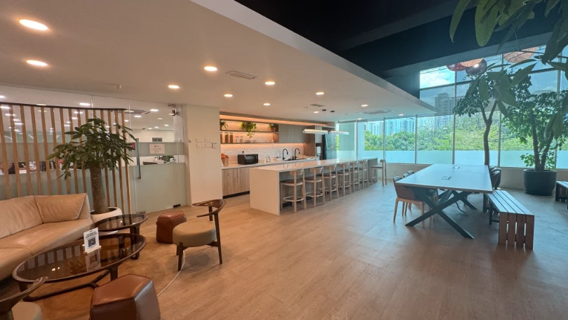 Modern open-plan office lounge and kitchen area with a long island and bar stools, a large table with chairs and bench, sofas, and large windows with city and tree views.