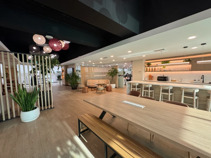 Modern office lounge area with wooden flooring, a large table with benches, a kitchen counter with stools, plants, and cozy seating.