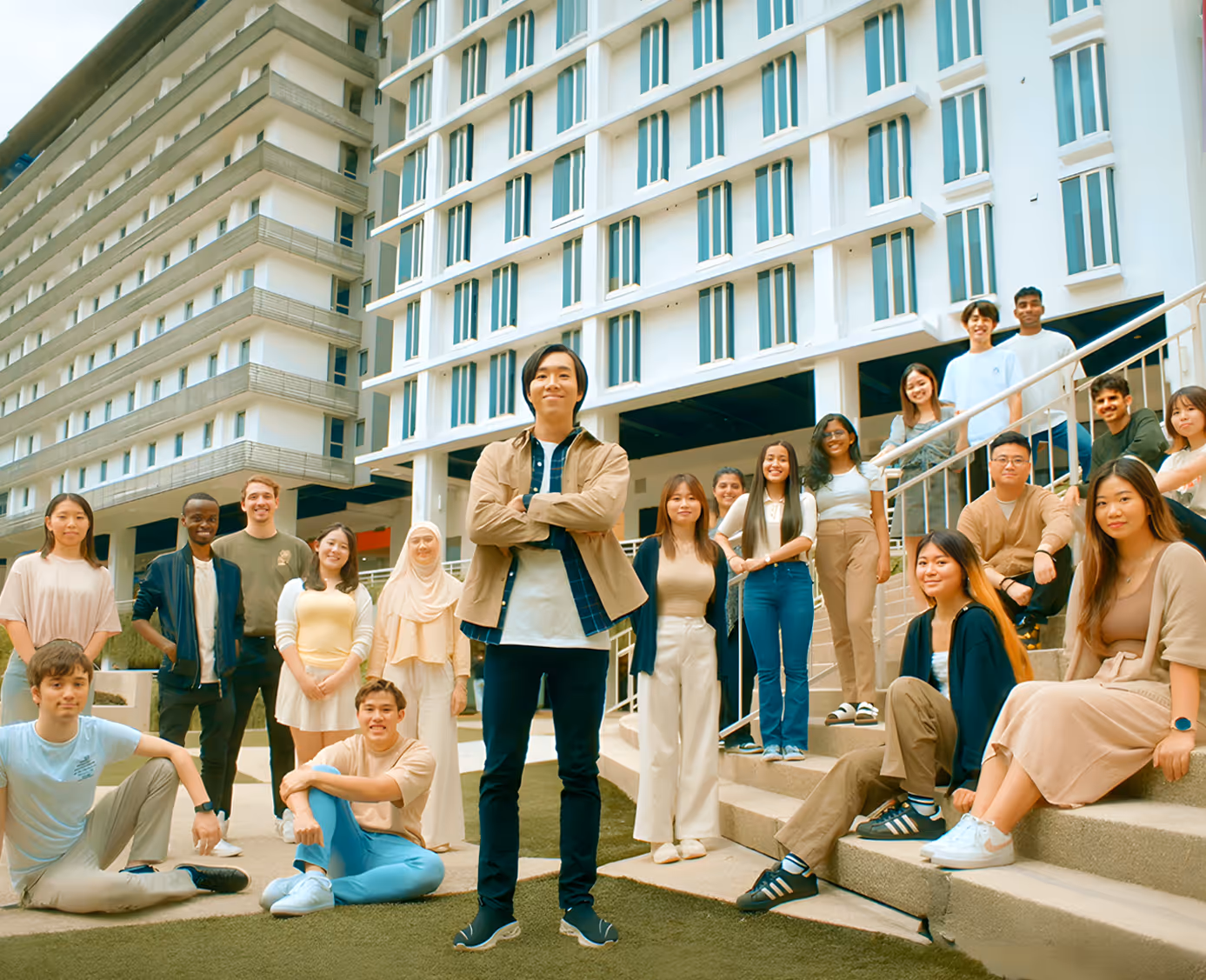 Diverse group of young adults posing outdoors in front of a modern building, some standing and others sitting on stairs.
