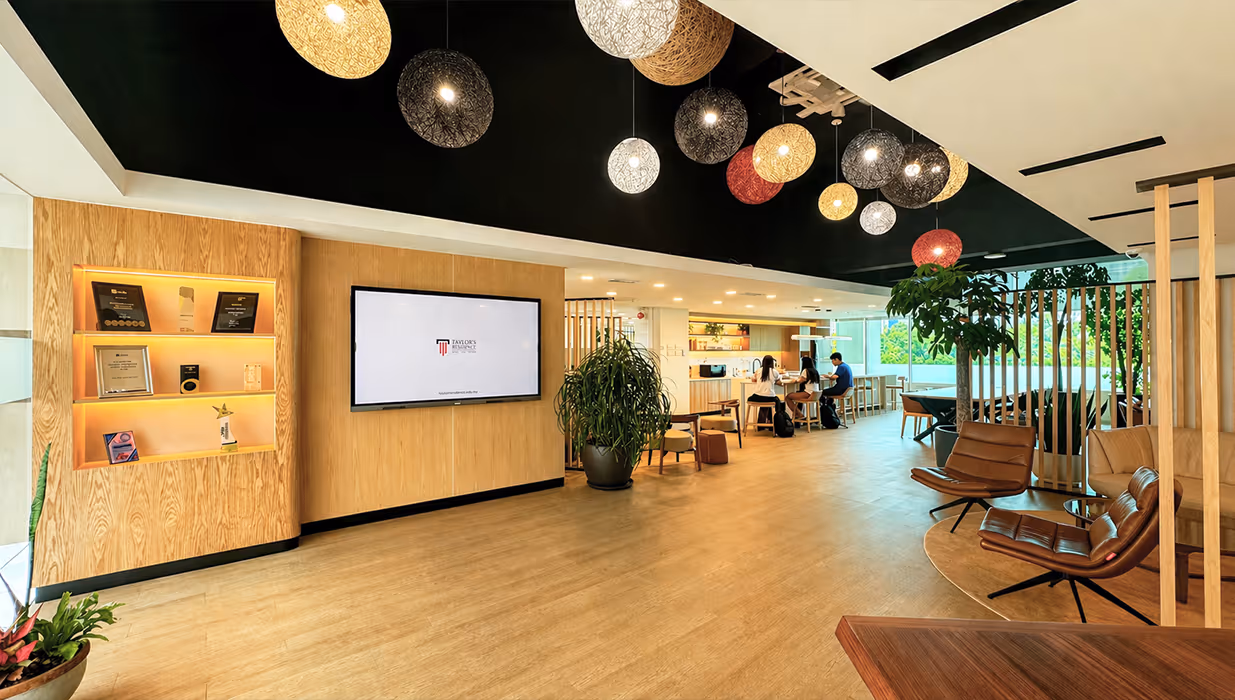 Modern office lobby with wooden floor and walls, hanging round pendant lights, a wall-mounted TV, a trophy shelf, indoor plants, and people sitting and working at a table.