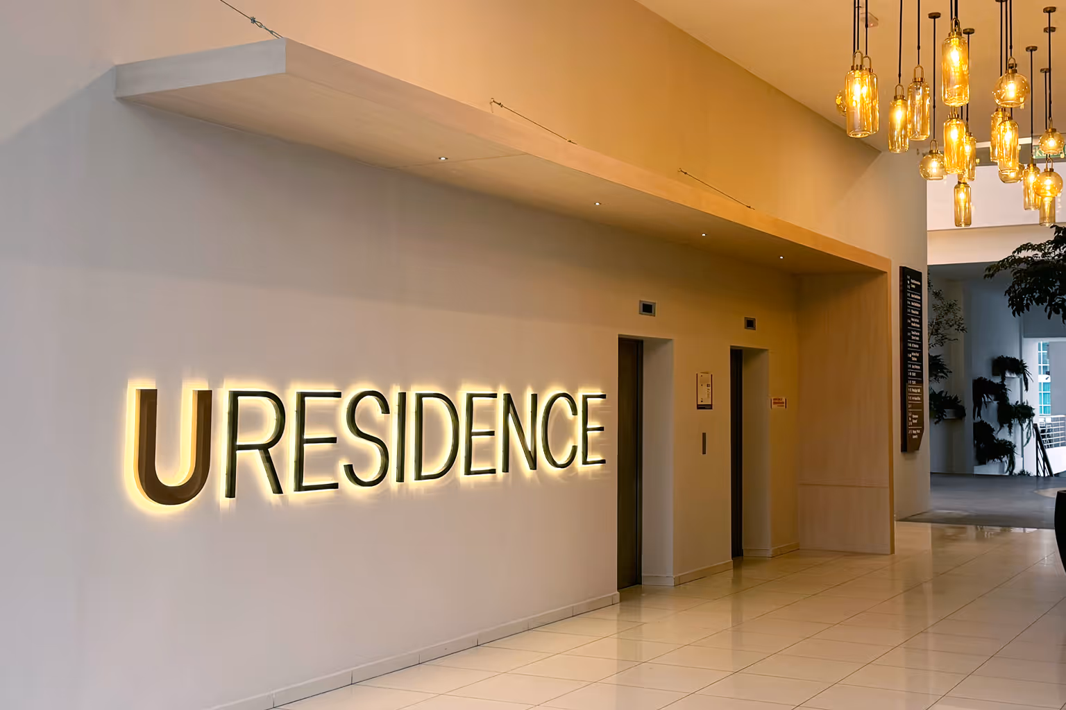 Modern lobby area with a glowing URESIDENCE sign on the wall, two elevator doors, and hanging pendant lights.