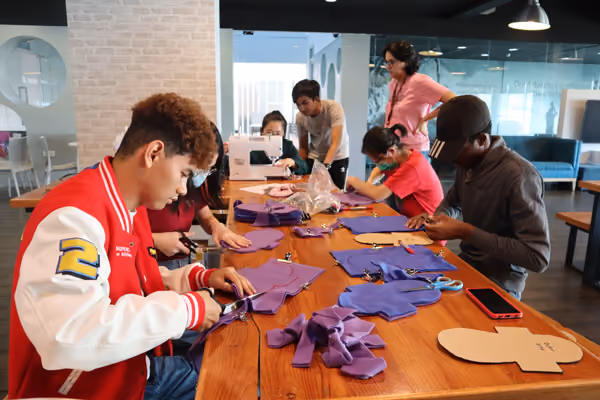 A group of young people sitting at a wooden table cutting purple fabric pieces using scissors, supervised by two adults in a modern room.