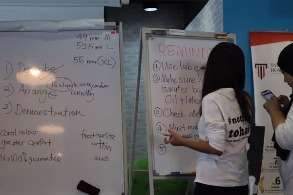 Two people standing next to whiteboards with handwritten notes about condom use and reminders in a classroom or workshop setting.
