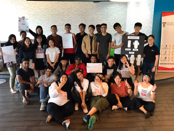 A diverse group of smiling young people posing indoors, some holding drawings and posters, in a casual meeting or workshop setting.