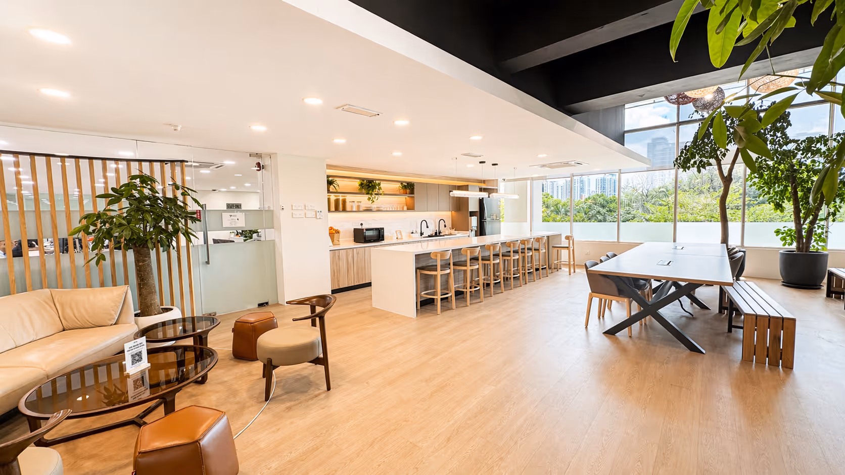 Modern office pantry with a long white island and stools, a dining table with chairs and benches, and a lounge area with sofa and chairs by large windows with greenery outside.