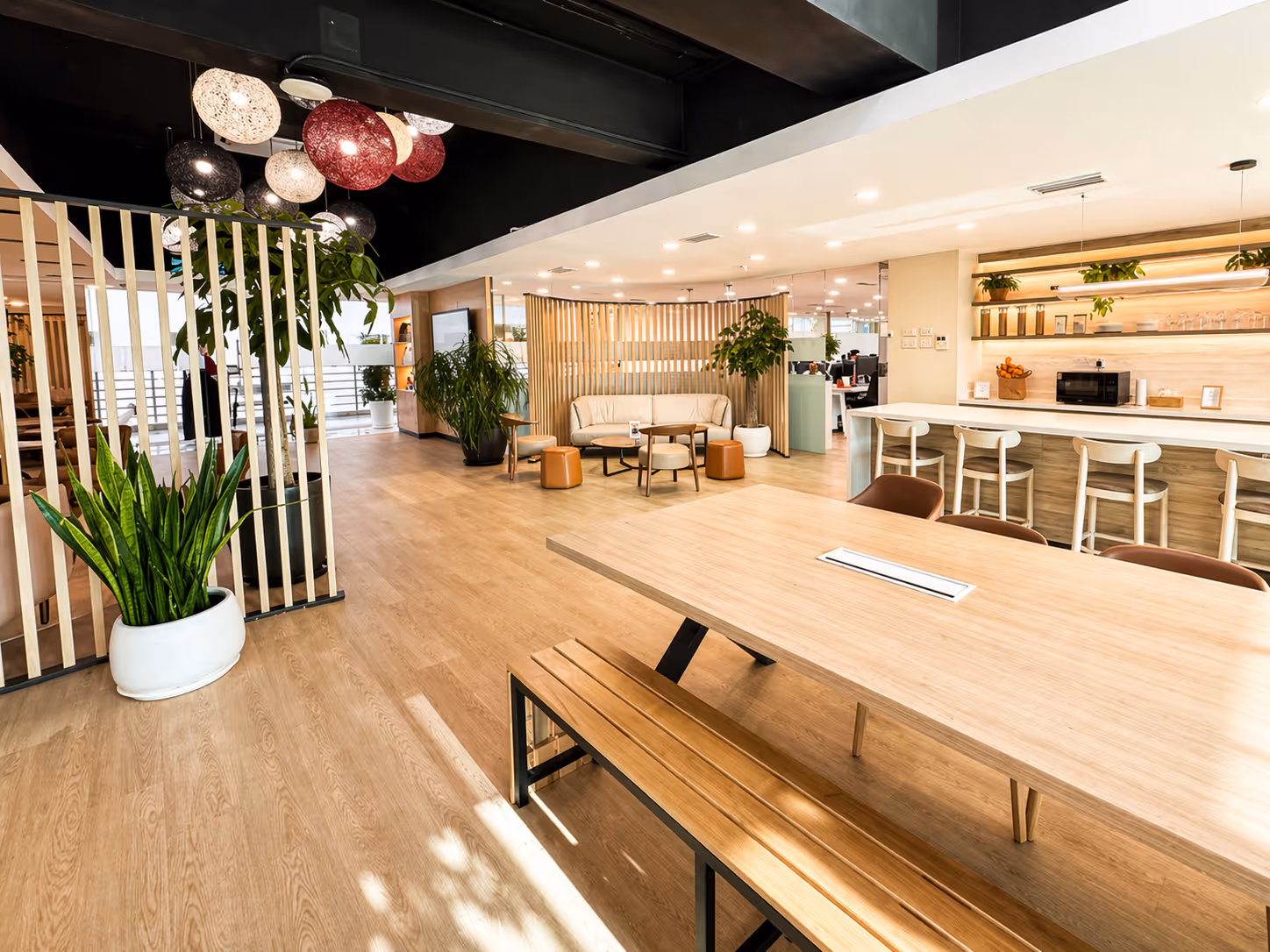Modern office lounge area with wooden floors, a long table with bench seating, a bar counter with stools, plants, and pendant lights.