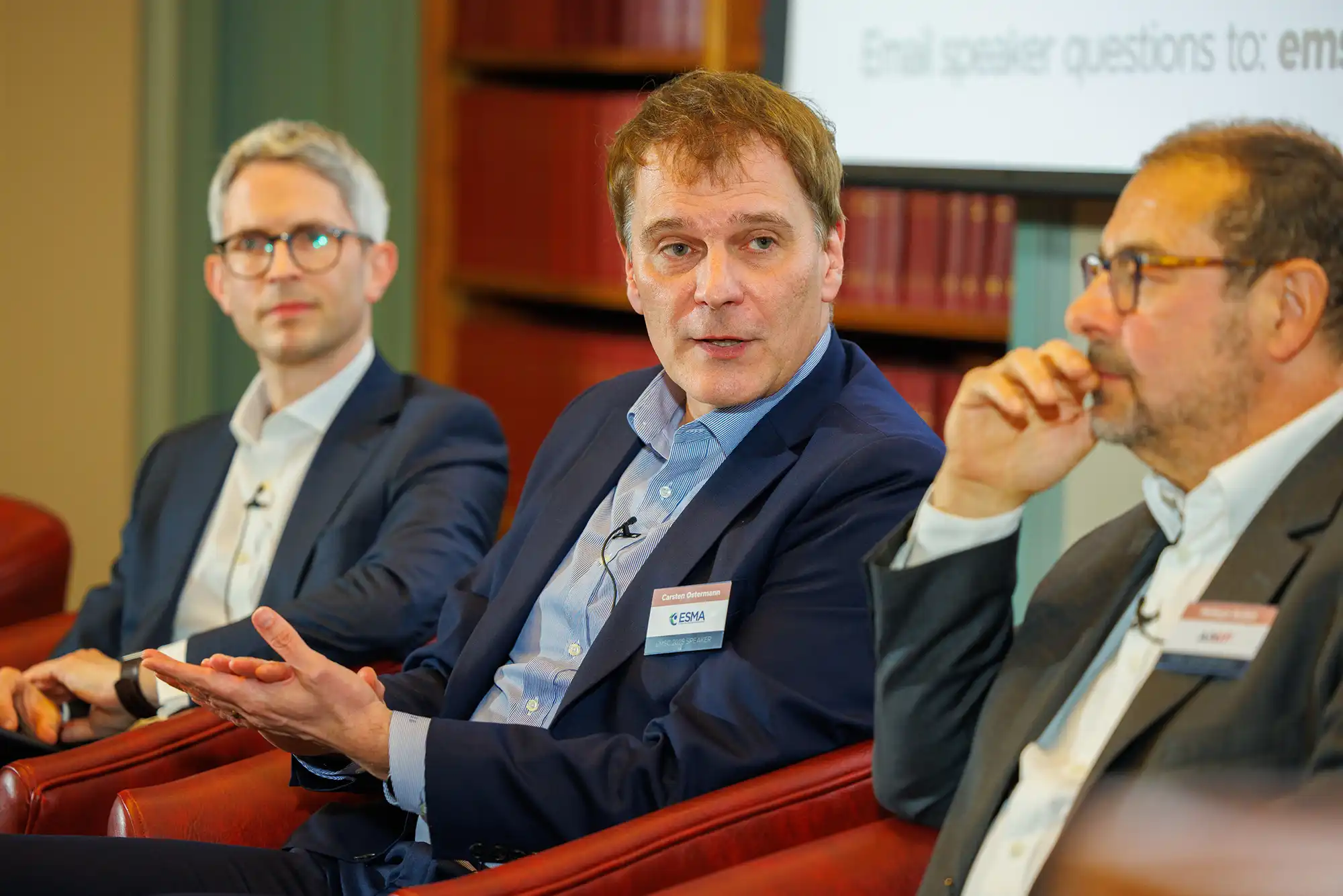 Carsten Ostermann from ESMA speaks on a panel at a professional event. He wears a blue suit and a name badge while gesturing during his presentation.