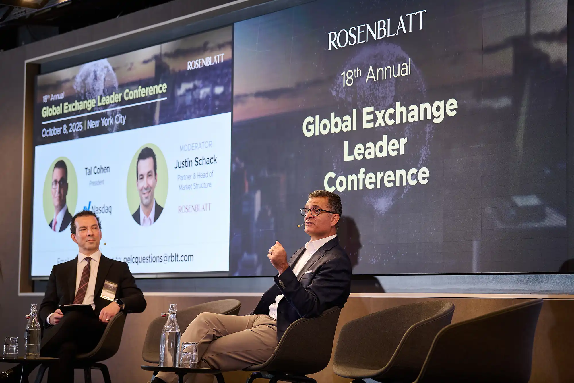 Moderator Justin Schack sits on stage while Nasdaq President Tal Cohen speaks at the Global Exchange Leader Conference.