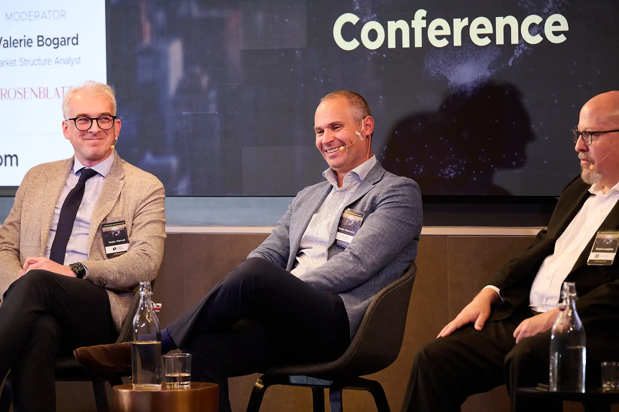 Three male panelists in business attire sit on stage and smile during a corporate discussion at an industry conference.