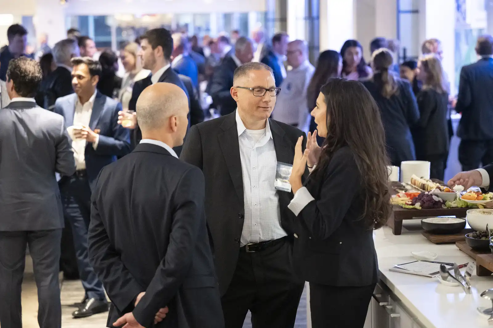 Three professionals chatting near a buffet table at a busy corporate networking event while other attendees mingle in the brightly lit background room.