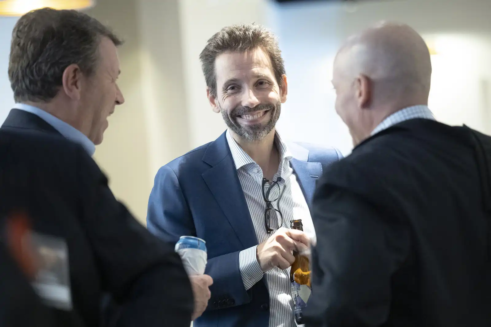 Three male colleagues sharing a laugh and holding drinks during a casual networking mixer, with the central man smiling warmly directly at his peers.