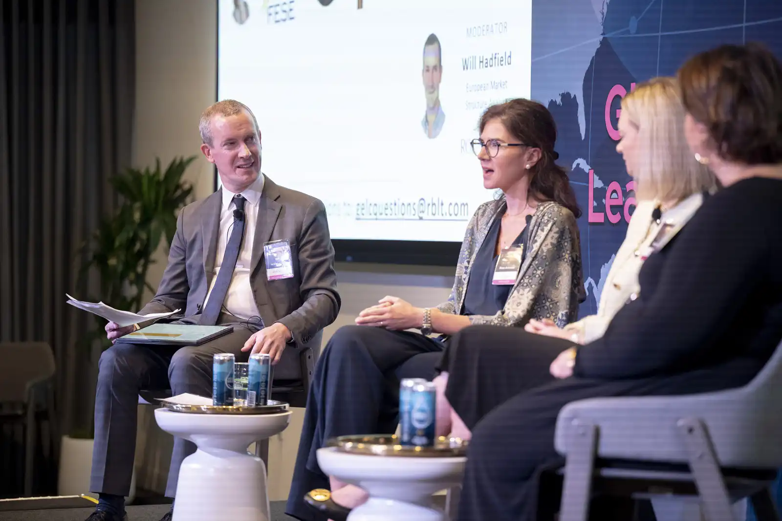 A diverse panel of four business experts participating in a lively stage discussion, seated on modern chairs before a large presentation screen.