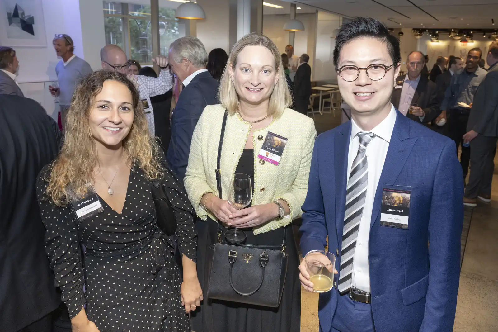 Three well-dressed conference attendees holding beverages and smiling for the camera while socializing together at an elegant indoor corporate reception.