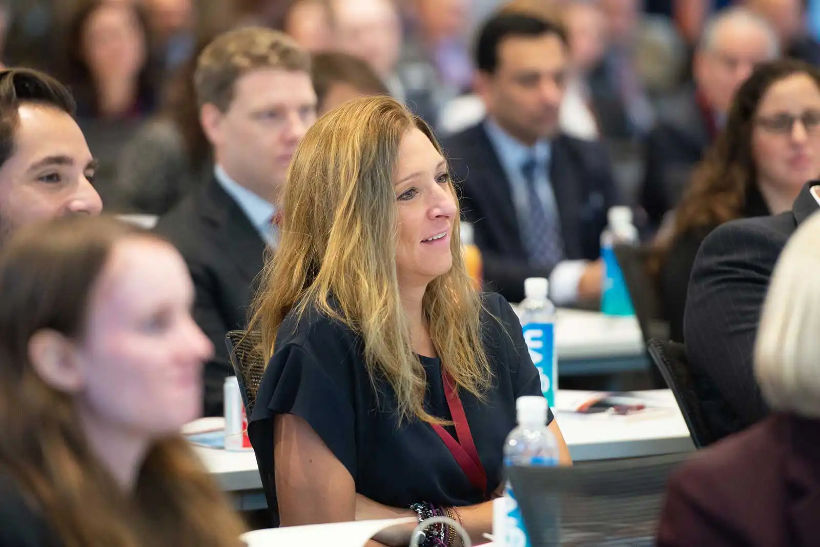 Engaged audience members seated at tables during a corporate presentation, focusing on a smiling blonde woman actively listening to the guest speaker.