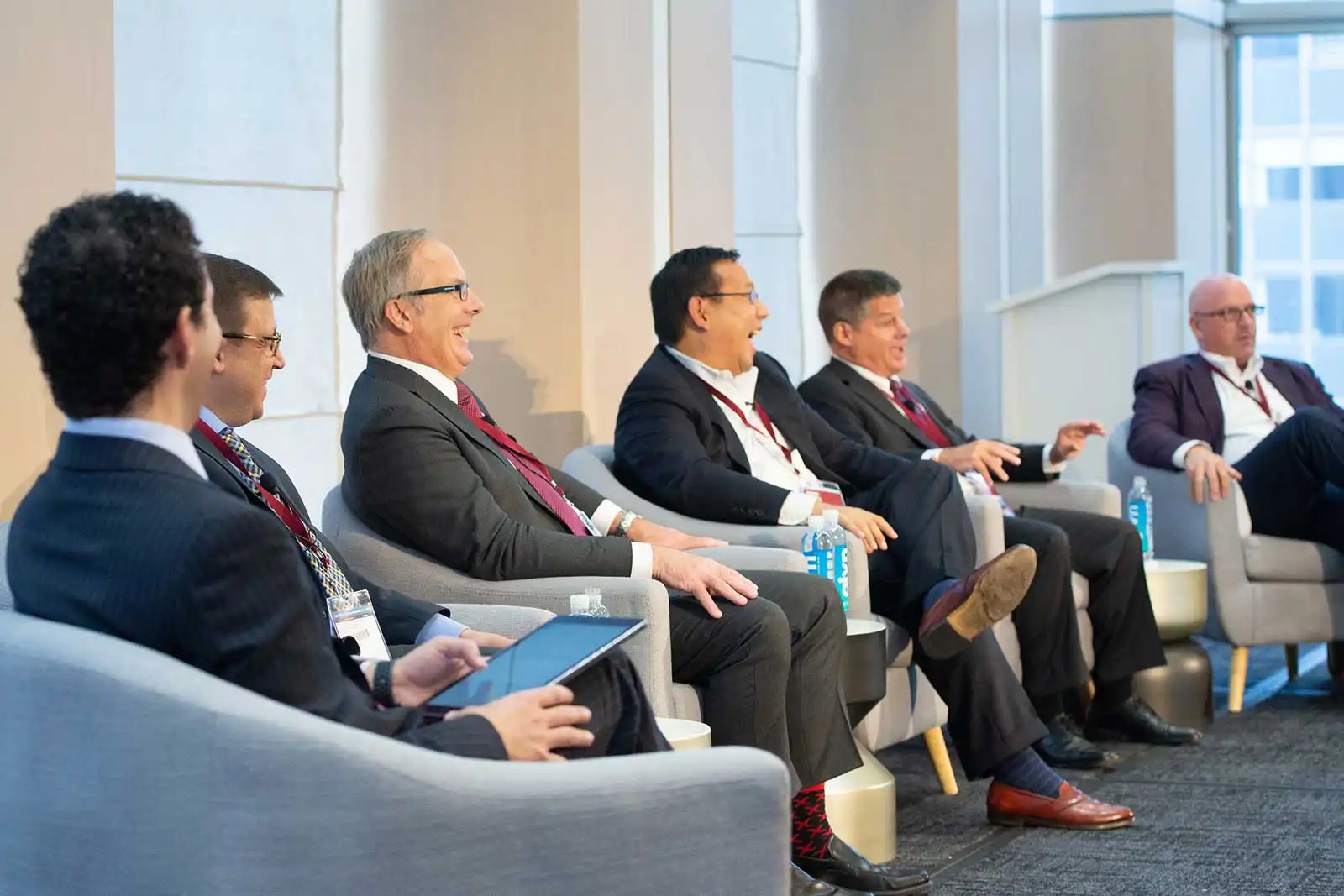 Five male business executives sitting in grey chairs on a brightly lit stage, sharing a humorous moment and laughing together during a panel discussion.