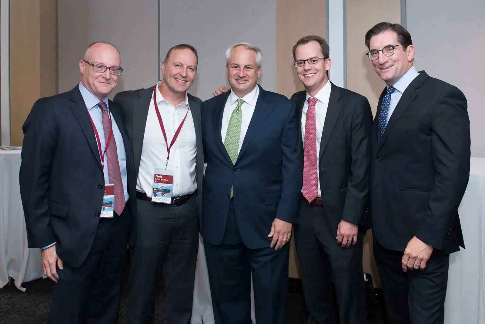 Five male business executives in professional suits smiling warmly while posing together for a group photo at a corporate industry conference.