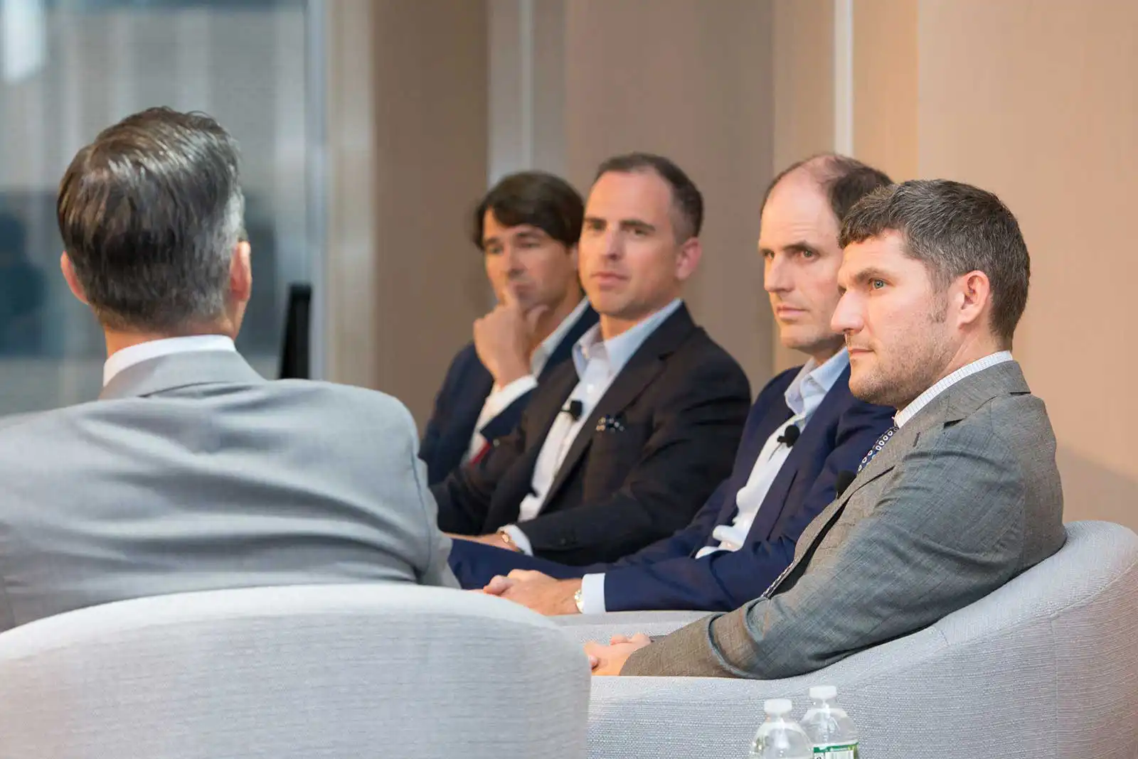 Four professional male panelists in business suits listening attentively during a serious stage discussion at an industry corporate conference.