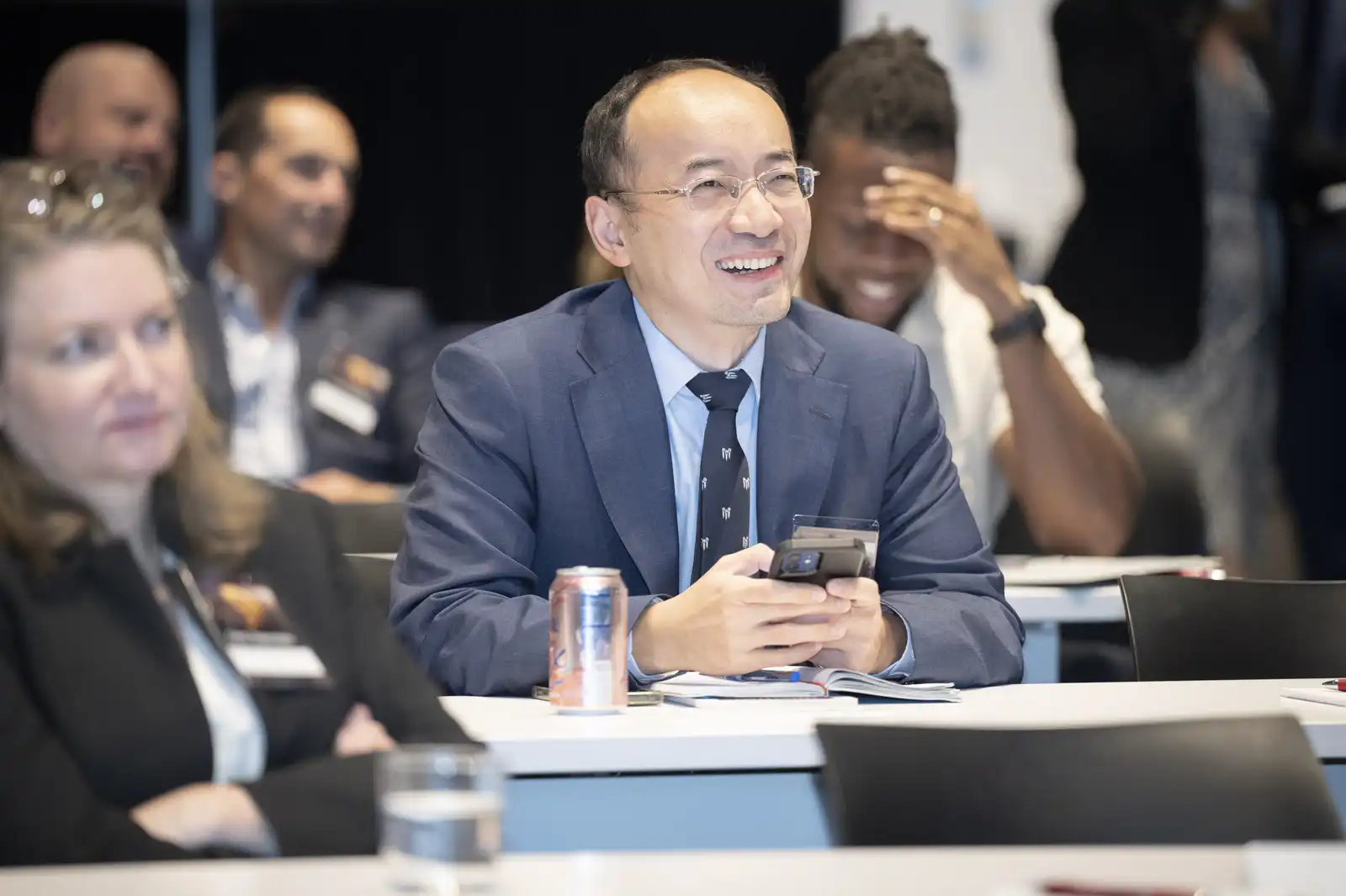 A cheerful male business professional wearing glasses and a blue suit smiling brightly while holding a smartphone at a corporate conference.