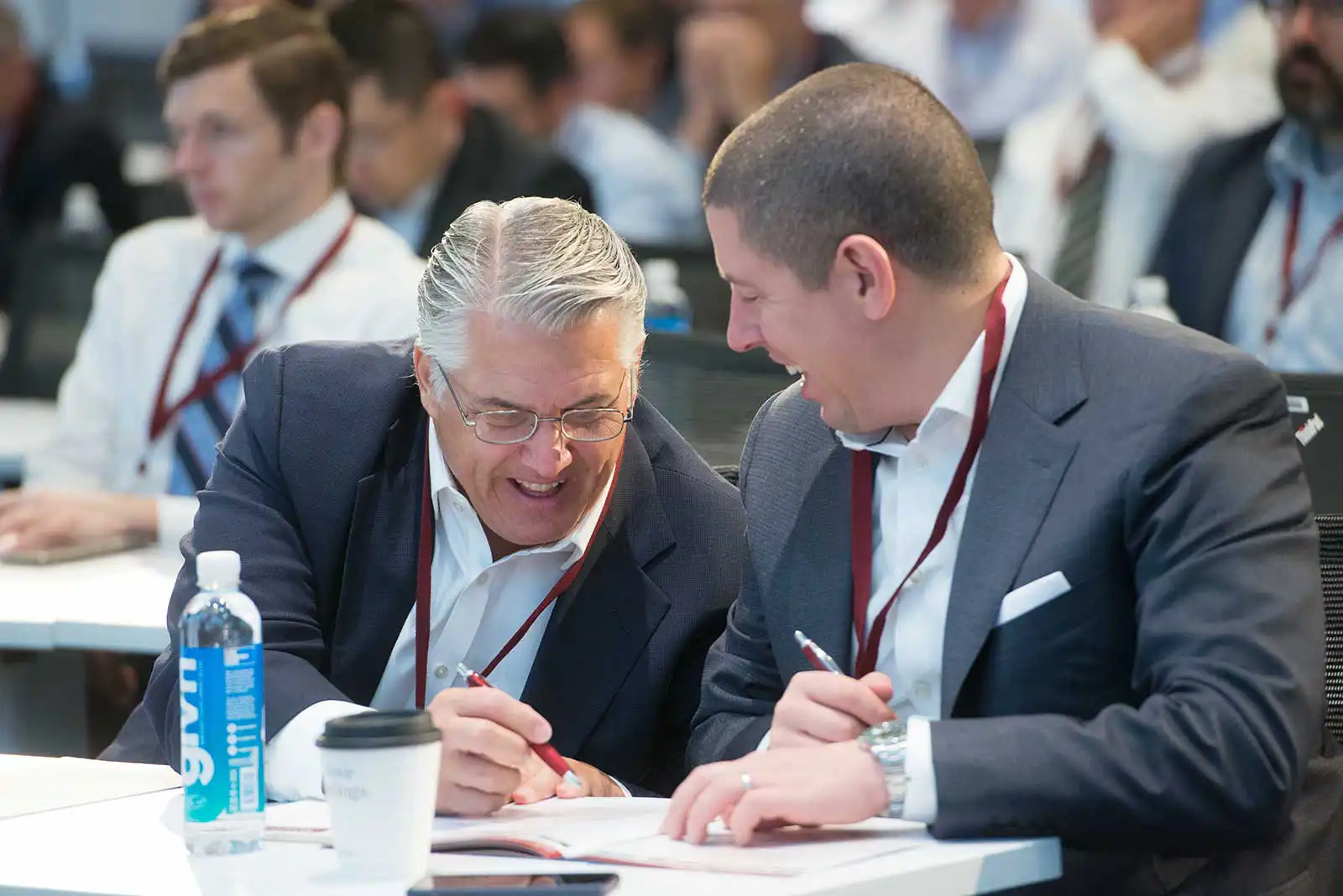 Two male corporate executives sharing a laugh while reviewing documents together at a table during a professional business industry conference.
