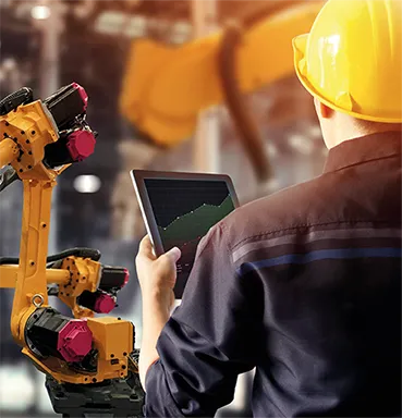 Ein Arbeiter mit gelbem Helm bedient ein Tablet vor einem gelben Industrieroboterarm in einer Fabrikumgebung.