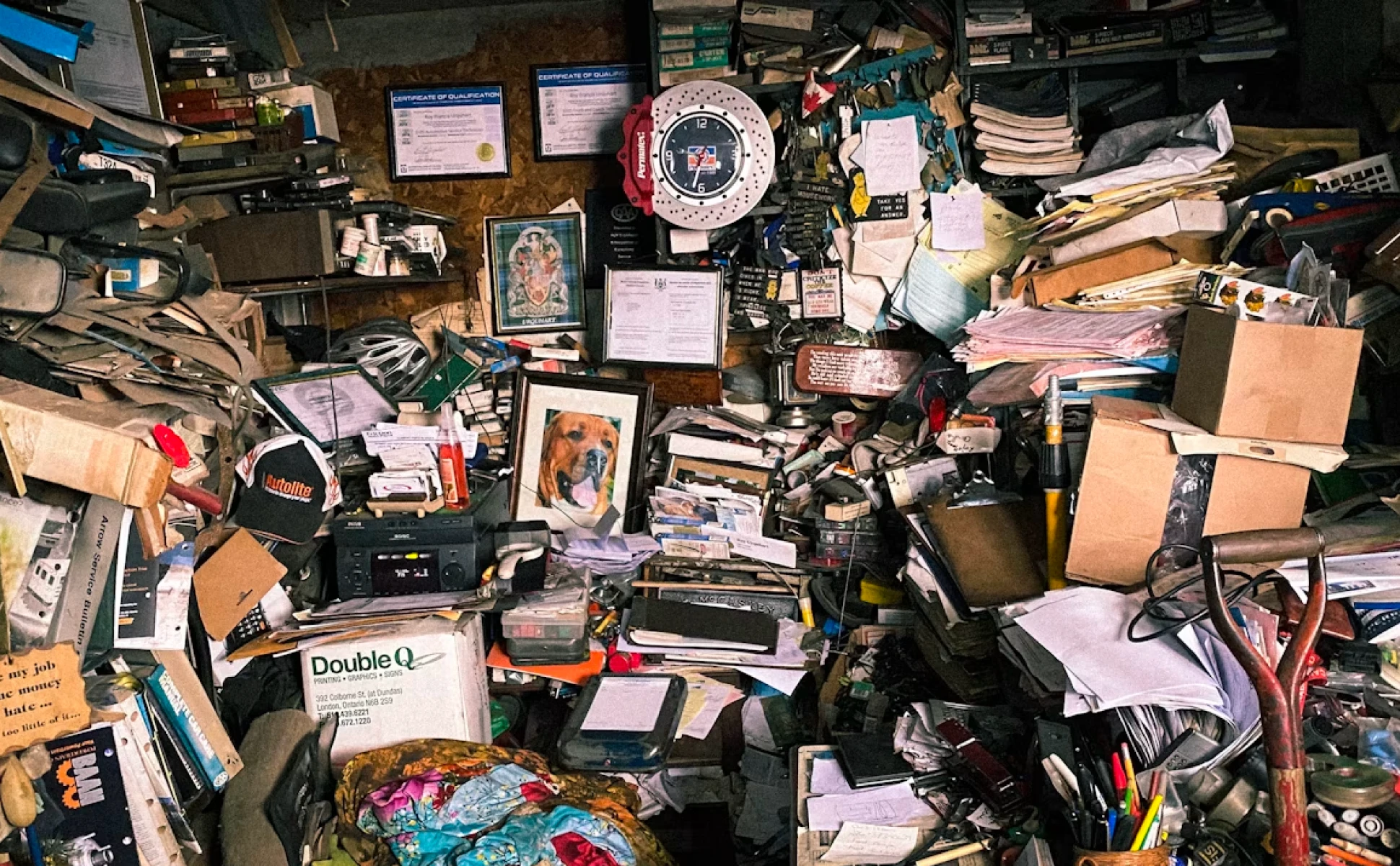 An extremely cluttered room and desk overflowing with stacked papers, cardboard boxes, certificates, framed photos, and dense visual noise.