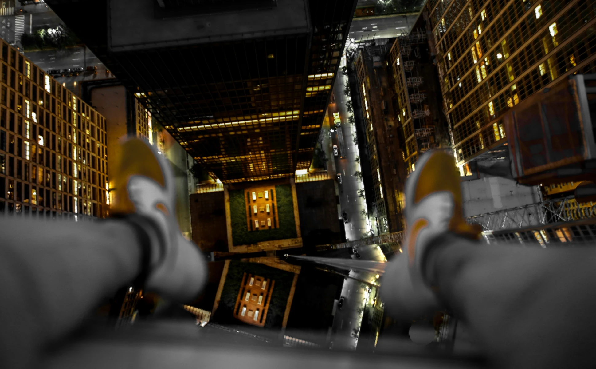 First-person POV looking straight down from the edge of a high-rise building at night, showing sneakers dangling over illuminated city streets and surrounding skyscrapers.