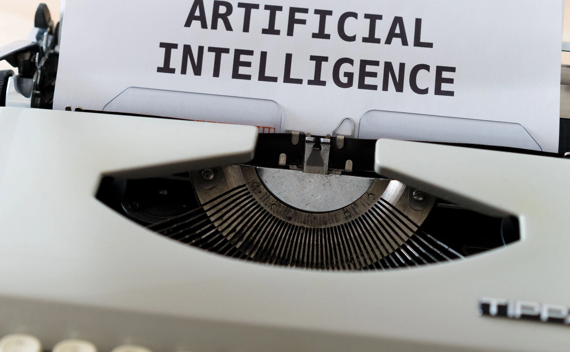 Close-up of a white vintage typewriter with a sheet of paper that has "ARTIFICIAL INTELLIGENCE" typed in bold, black capital letters.