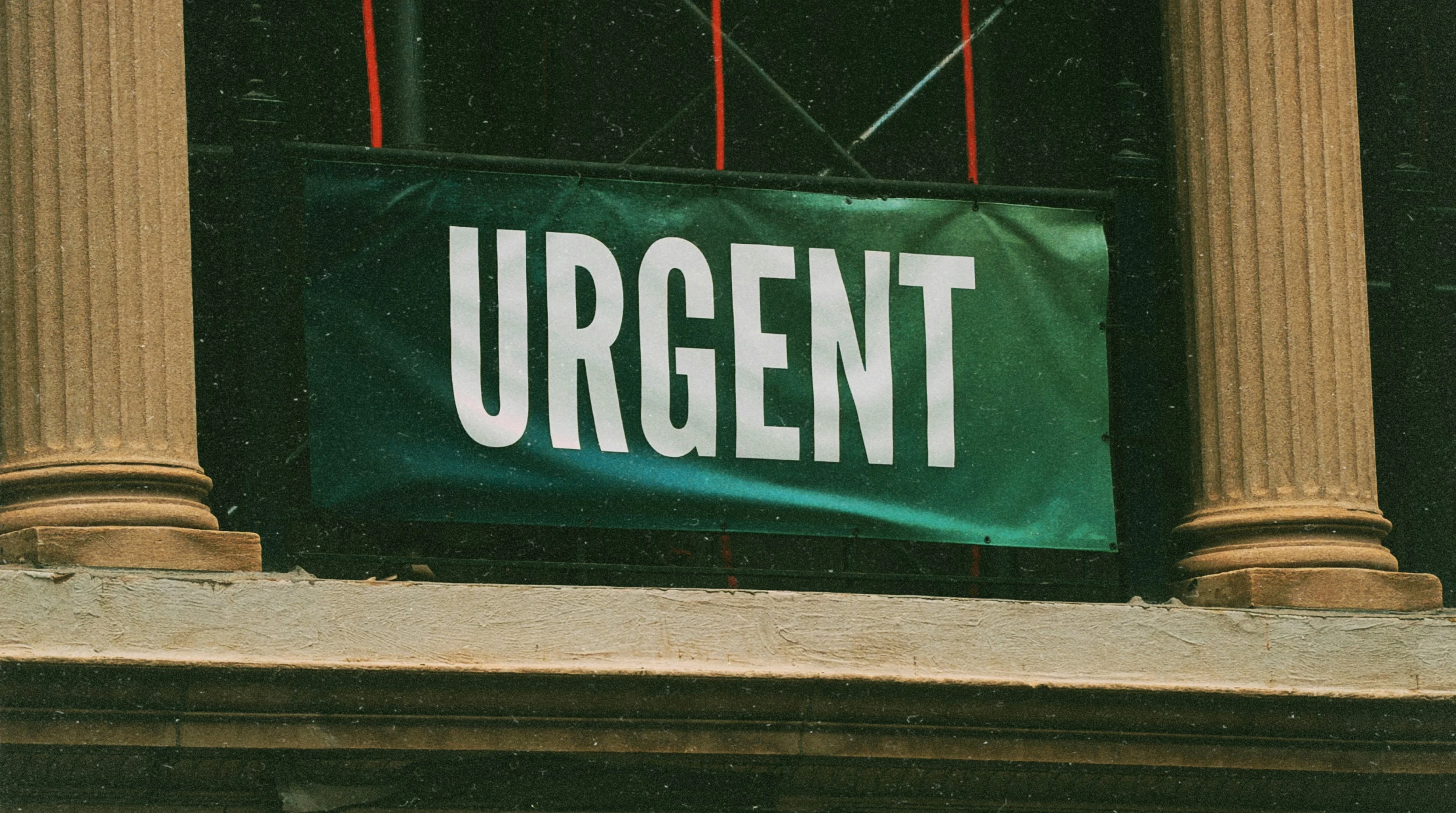 Green banner reading URGENT in bold white capitals hanging between two stone columns on an old building, with red cords and scaffolding behind.