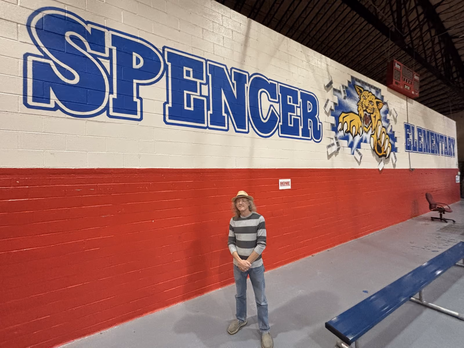 Local artist Steve Adams stands in front of his completed mural in the Spencer Elementary School gymnasium near the end of the 2025–2026 school year. The massive wall display features bold lettering and the SES Wildcat mascot bursting through the brickwork