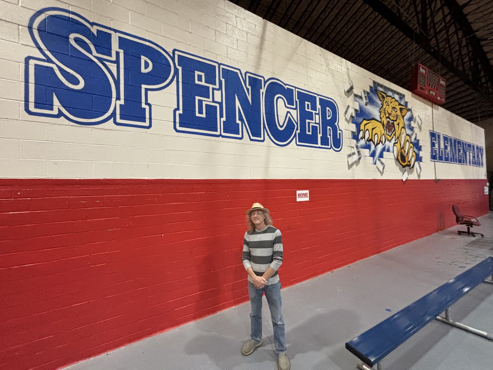Local artist Steve Adams stands in front of his completed mural in the Spencer Elementary School gymnasium near the end of the 2025–2026 school year. The massive wall display features bold lettering and the SES Wildcat mascot bursting through the brickwork