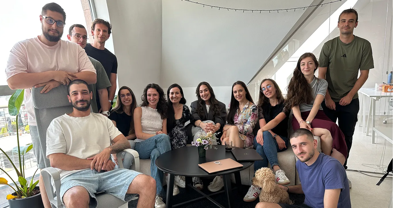 Group of 13 young adults smiling in an office lounge around a black table with laptops and a small flower vase, with one person holding a small dog.