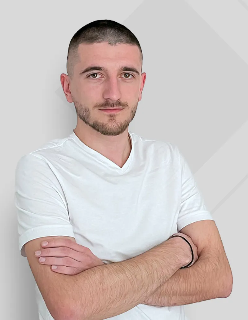 Young man with short hair and beard wearing a white t-shirt, arms crossed, against a light gray background.