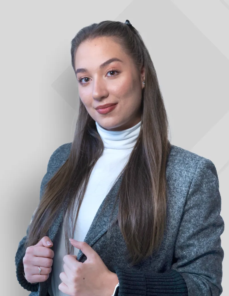 Young woman with long brown hair wearing a gray blazer over a white turtleneck, smiling gently against a light gray background.