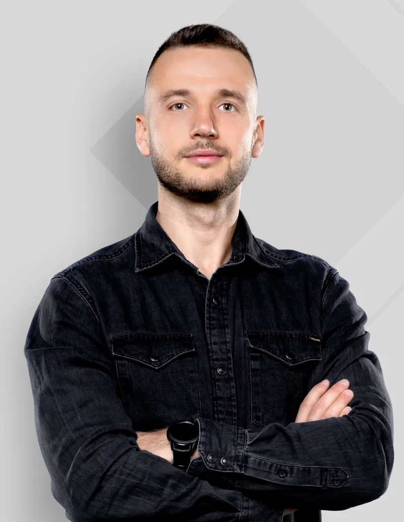 Confident young man with short hair and beard wearing a black denim shirt and smartwatch, arms crossed.