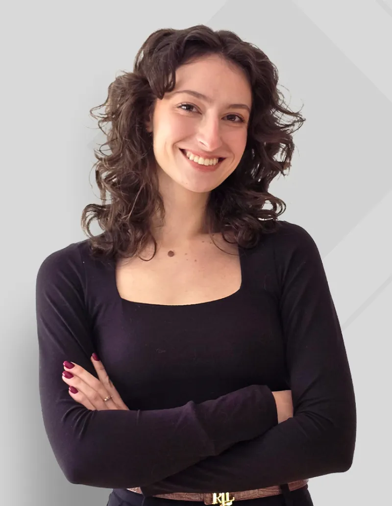 Smiling woman with curly brown hair wearing a black long-sleeve top, standing with arms crossed against a light gray background.